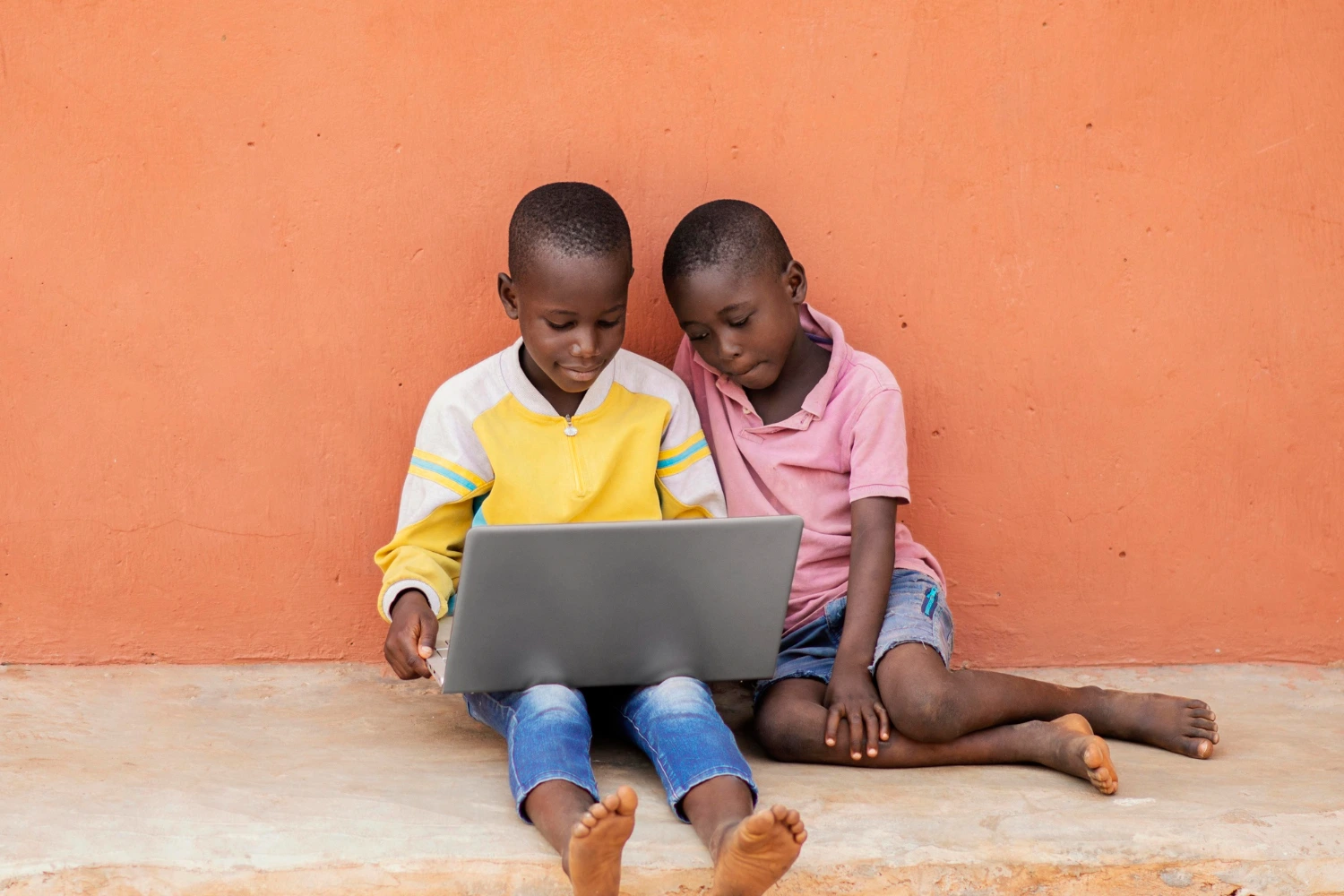 Two young boys seated on the ground, focused on a laptop in front of them, engaged in a shared activity.