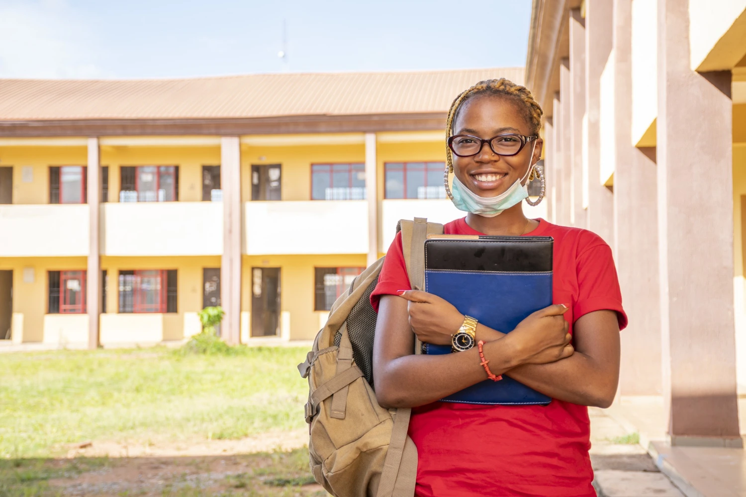 A young woman wearing a face mask and glasses stands outside a school building, looking towards the camera.